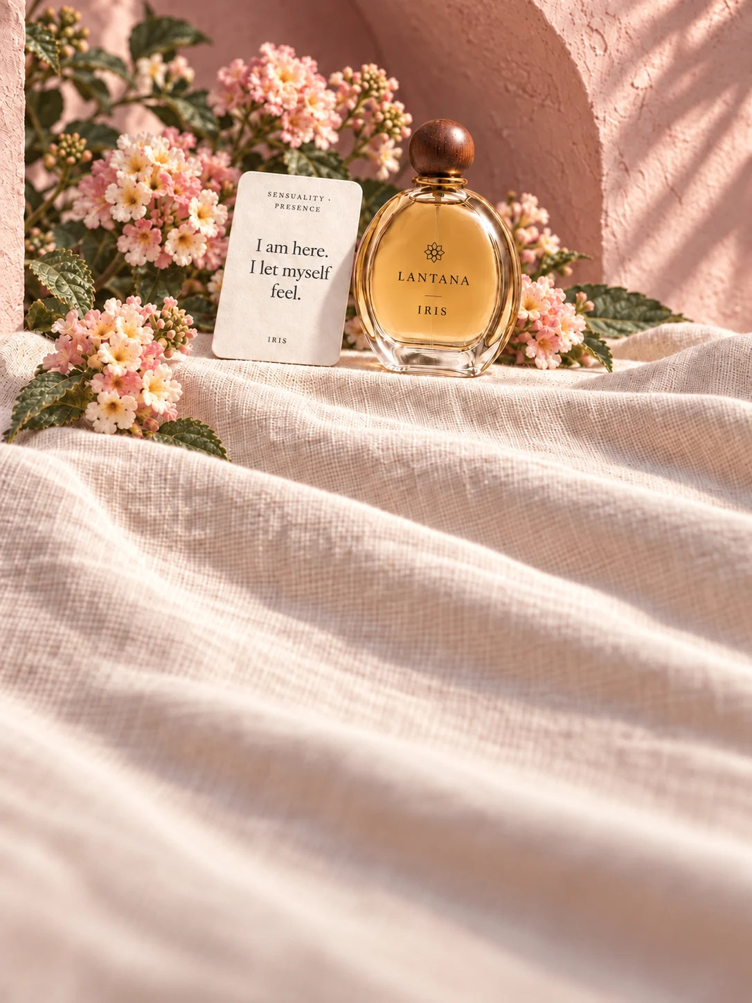 IRIS perfume bottle and its Sensuality and Presence intention card with blush lantana and softly draped ivory linen in a pink plaster alcove.
